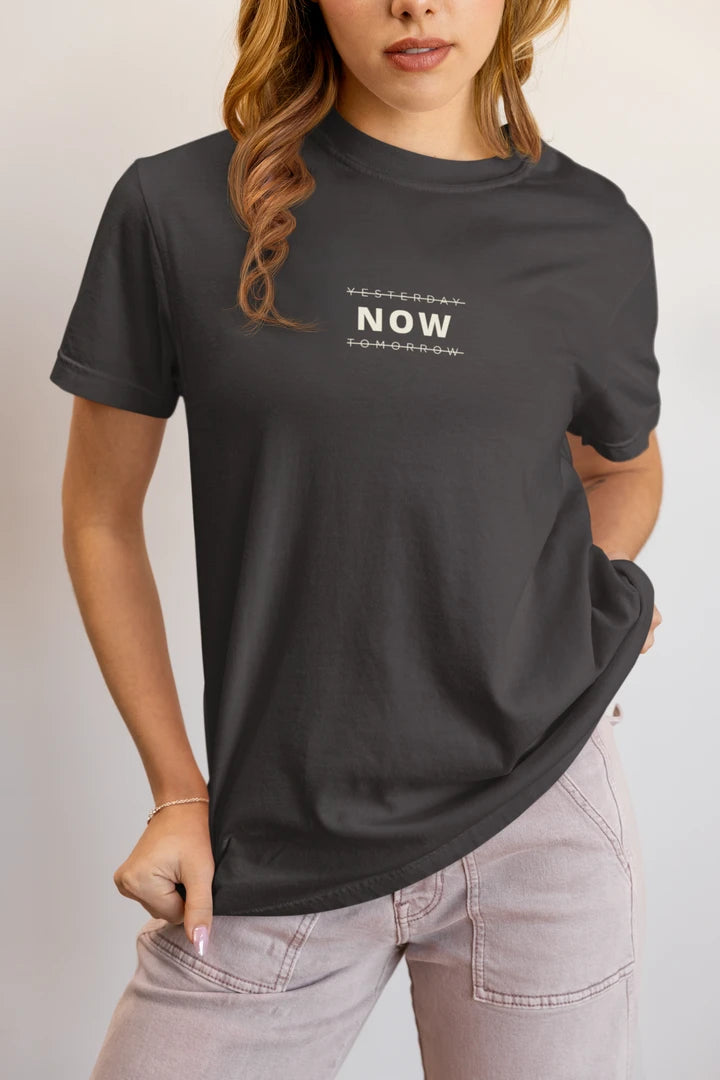 A woman wearing a Graphite t-shirt with a white 'YESTERDAY NOW TOMORROW' logo on the chest, paired with light pink jeans, against a plain light gray background.