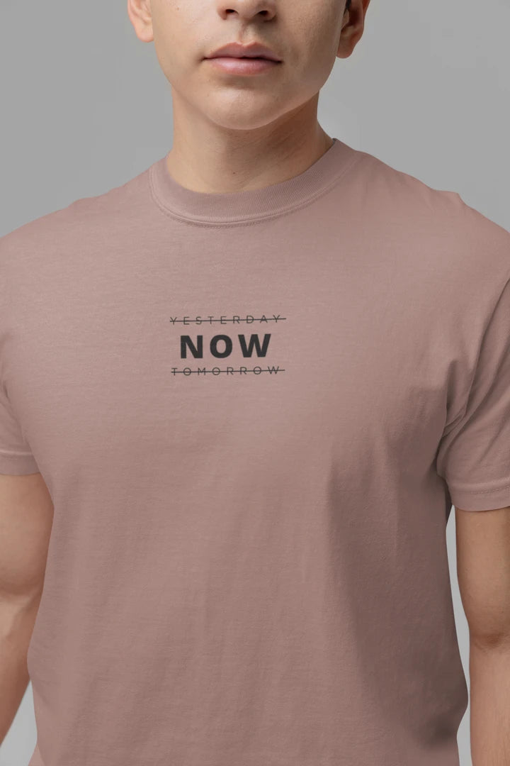 A close-up of a person wearing a taupe t-shirt with a black graphic text print reading 'YESTERDAY NOW TOMORROW' on the chest, set against a solid gray background.