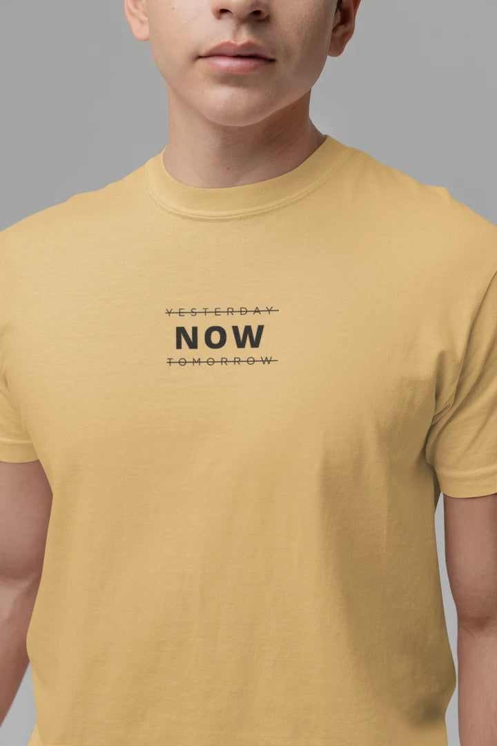 A close-up of a person wearing a Warm sand t-shirt with a black graphic text print reading 'YESTERDAY NOW TOMORROW' on the chest, set against a solid gray background.