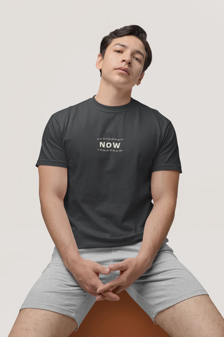 A man wearing a Graphite t-shirt with a white 'Yesterday NOW Tomorrow' logo on the chest, paired with light gray shorts, sitting against a light beige background.