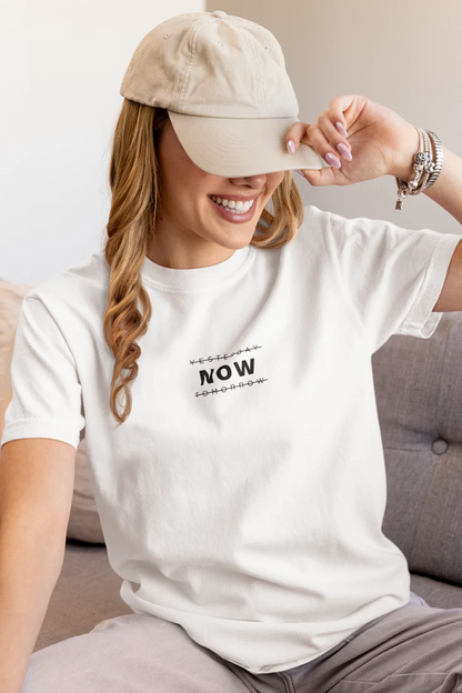 Woman wearing a Pure white t-shirt with a 'YESTERDAY NOW TOMORROW' graphic and a beige cap, sitting on a couch.
