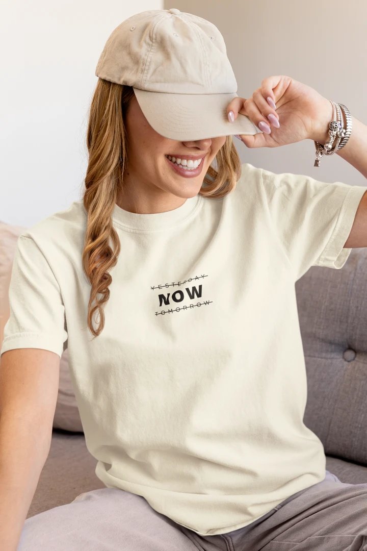 A woman wearing a off white t-shirt with a black graphic print that says 'YESTERDAY NOW TOMORROW' and a beige baseball cap, sitting on a gray sofa.