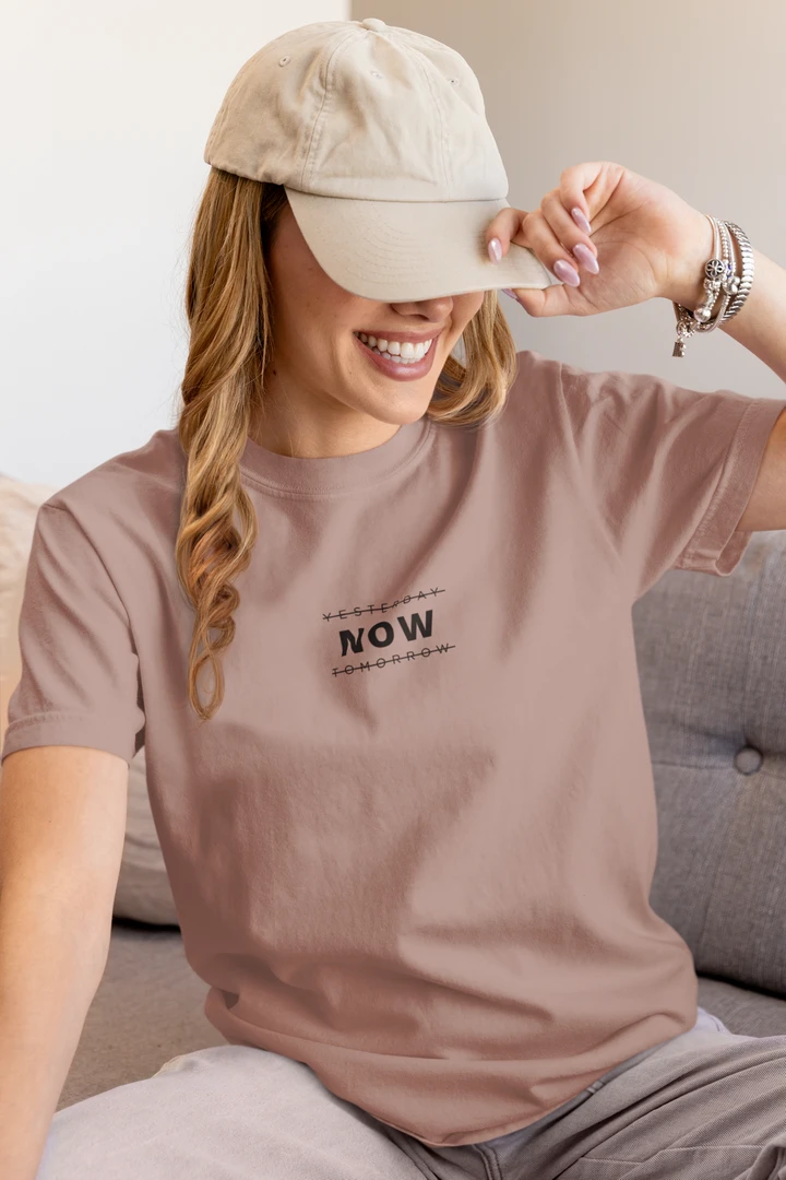 A woman wearing a taupe t-shirt with a black graphic print that reads 'YESTERDAY NOW TOMORROW' and a beige baseball cap, sitting on a gray sofa.