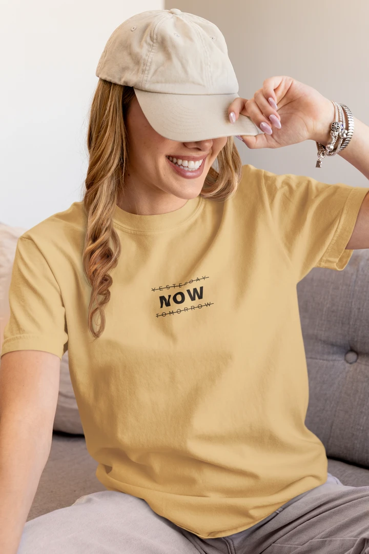 A woman sitting on a gray sofa wearing a Warm sand t-shirt with a black graphic print that says 'YESTERDAY NOW TOMORROW' and a beige baseball cap.