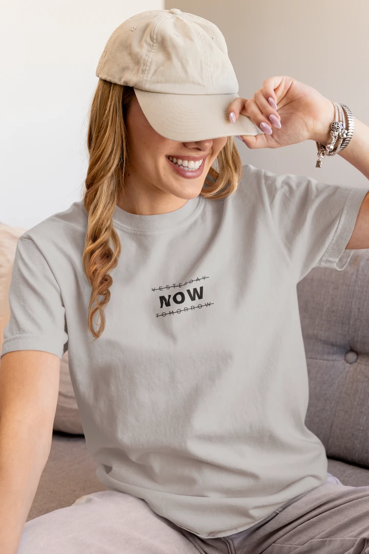 A woman wearing a heather grey t-shirt with a black graphic print that says 'YESTERDAY NOW TOMORROW' and a beige baseball cap, sitting on a gray sofa.
