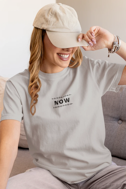 A woman wearing a heather grey t-shirt with a black graphic print that says 'YESTERDAY NOW TOMORROW' and a beige baseball cap, sitting on a gray sofa.