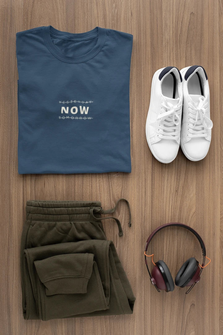 A styled flat-lay outfit on a wood floor featuring a Midnight Navy t-shirt with white text, olive green sweatpants, white sneakers, and black over-ear headphones.