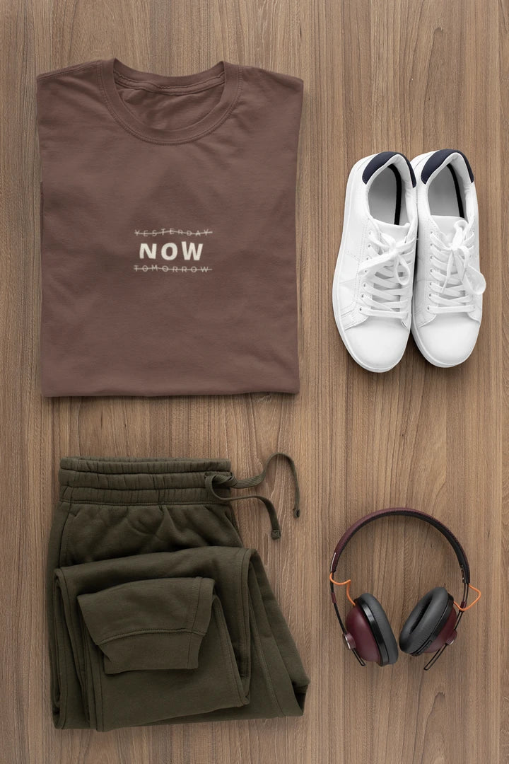 A styled flat-lay of a Dark Mocha t-shirt with white sneakers, olive green sweatpants, and over-ear headphones on a light brown wood background. The t-shirt features the word 'NOW' printed in white on the chest.