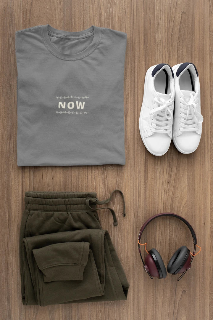 A styled flat-lay outfit on a wood background featuring a folded heather charcoal t-shirt with 'YESTERDAY NOW TOMORROW' printed on the chest, olive green sweatpants, white sneakers, and over-ear headphones.