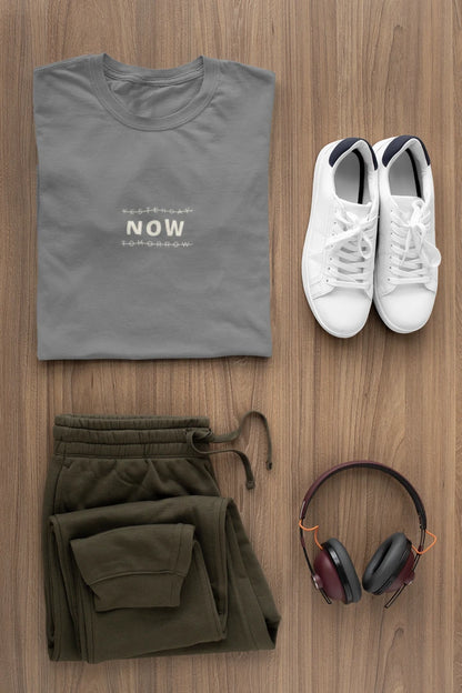 A styled flat-lay outfit on a wood background featuring a folded heather charcoal t-shirt with 'YESTERDAY NOW TOMORROW' printed on the chest, olive green sweatpants, white sneakers, and over-ear headphones.