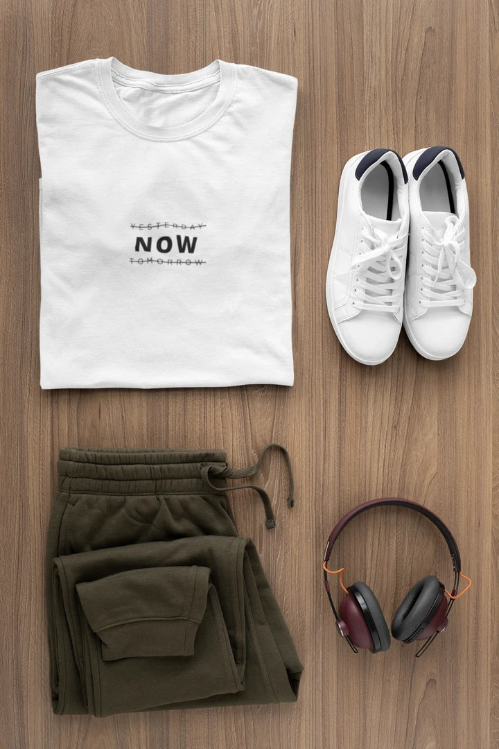 A styled flat-lay outfit on a wood floor featuring a Pure white t-shirt with black text, olive green sweatpants, white sneakers, and over-ear headphones.