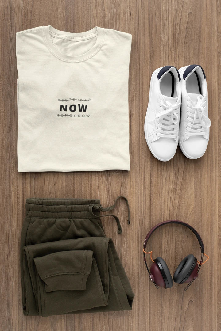 A styled flat-lay of a off white t-shirt with 'YESTERDAY NOW TOMORROW' printed on the front, paired with olive green sweatpants, white sneakers, and over-ear headphones on a wooden background.