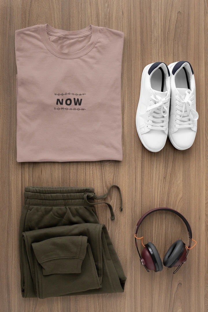 A styled flat-lay of a travel outfit on a wooden background, featuring a taupe t-shirt with the text 'YESTERDAY NOW Tomorrow', olive green sweatpants, white sneakers, and over-ear headphones.