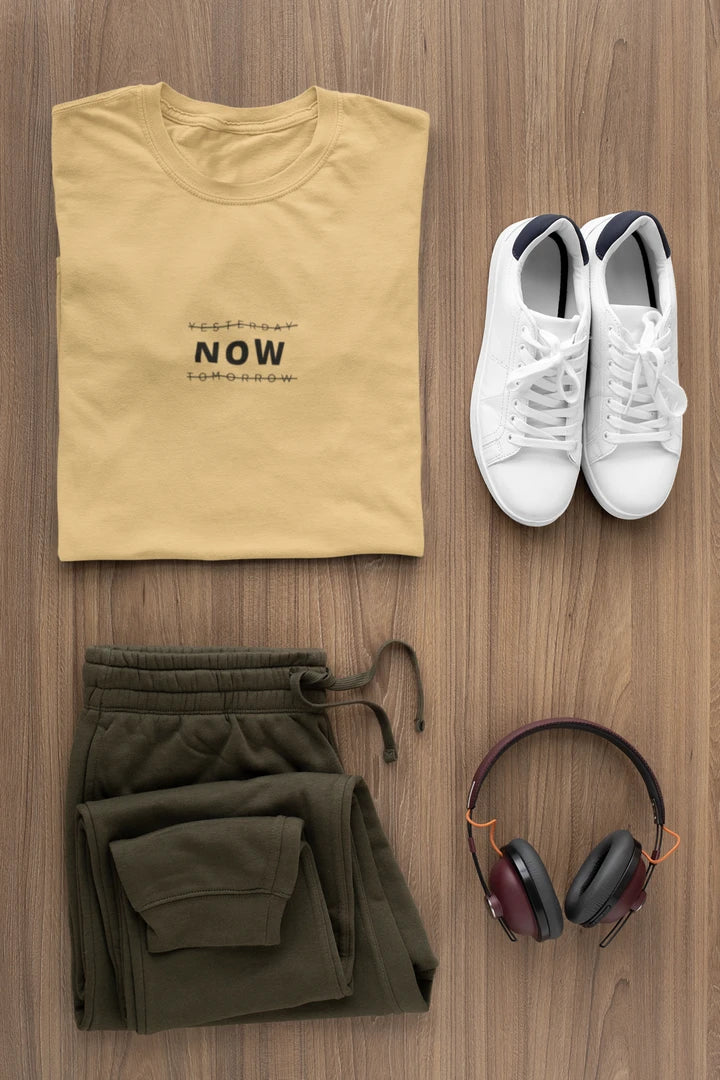 A styled flat-lay of a folded Warm sand t-shirt with 'YESTERDAY NOW TOMORROW' printed on the chest, olive green sweatpants, a pair of white sneakers, and over-ear headphones, arranged on a brown wood grain background.