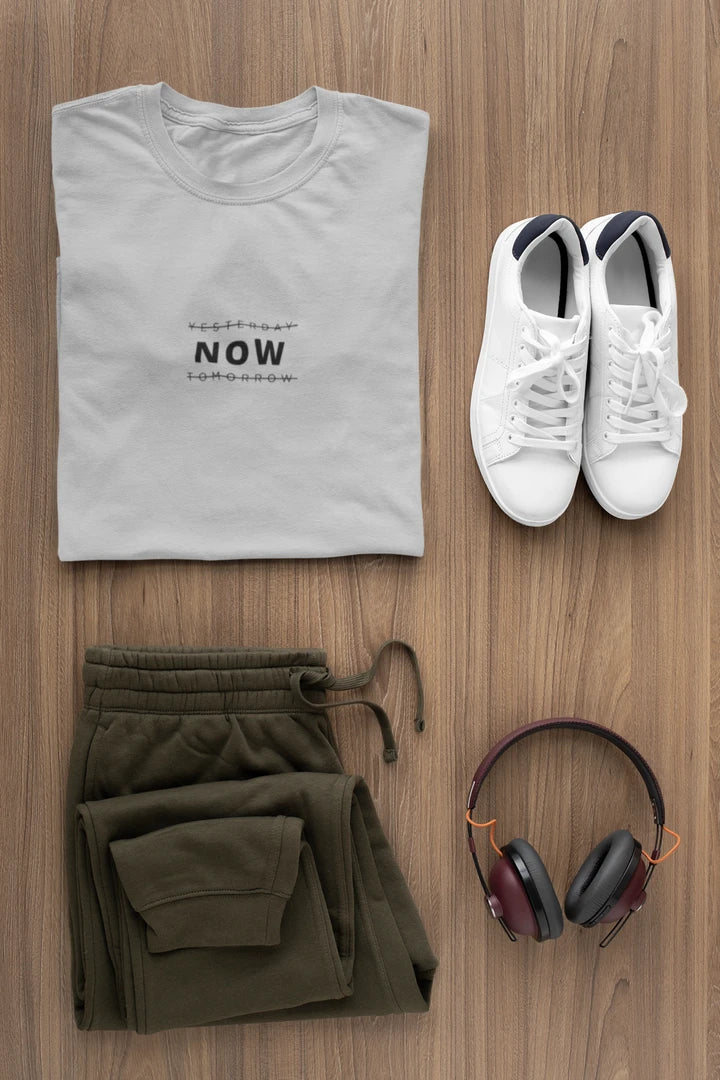 A styled flat-lay of a heather grey t-shirt with 'YESTERDAY NOW TOMORROW' printed on the chest, paired with olive green sweatpants, white sneakers, and over-ear headphones on a wooden background.