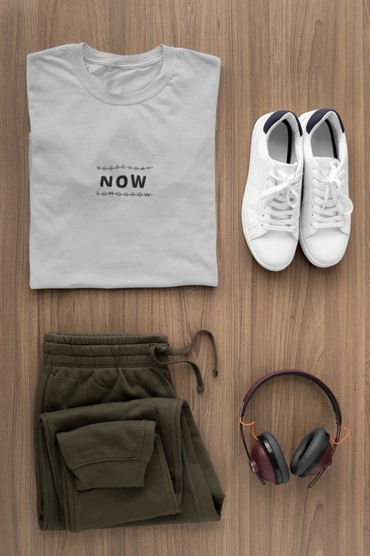 A styled flat-lay of a heather grey t-shirt with 'YESTERDAY NOW TOMORROW' printed on the chest, paired with olive green sweatpants, white sneakers, and over-ear headphones on a wooden background.