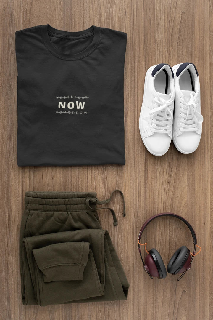 A styled flat-lay outfit on a wood background featuring a Jet Black t-shirt with white text, olive green sweatpants, white sneakers, and over-ear headphones.