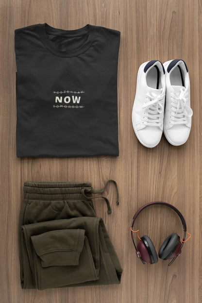 A styled flat-lay outfit on a wood background featuring a Jet Black t-shirt with white text, olive green sweatpants, white sneakers, and over-ear headphones.