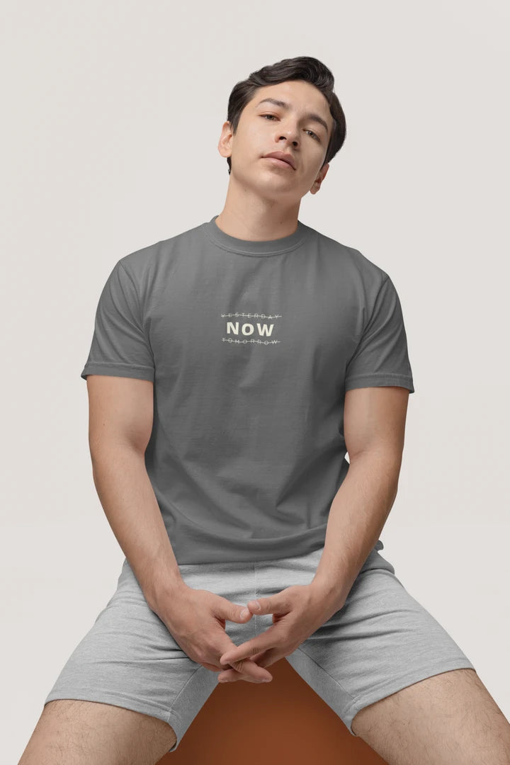 A man wearing a heather charcoal t-shirt with the text 'YESTERDAY NOW TOMORROW' printed in white on the chest, paired with light gray shorts, seated against a solid light gray background.