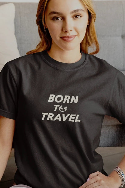 A woman wearing a Graphite t-shirt with the text 'BORN TO TRAVEL' printed in white on the chest, sitting in front of a gray couch.