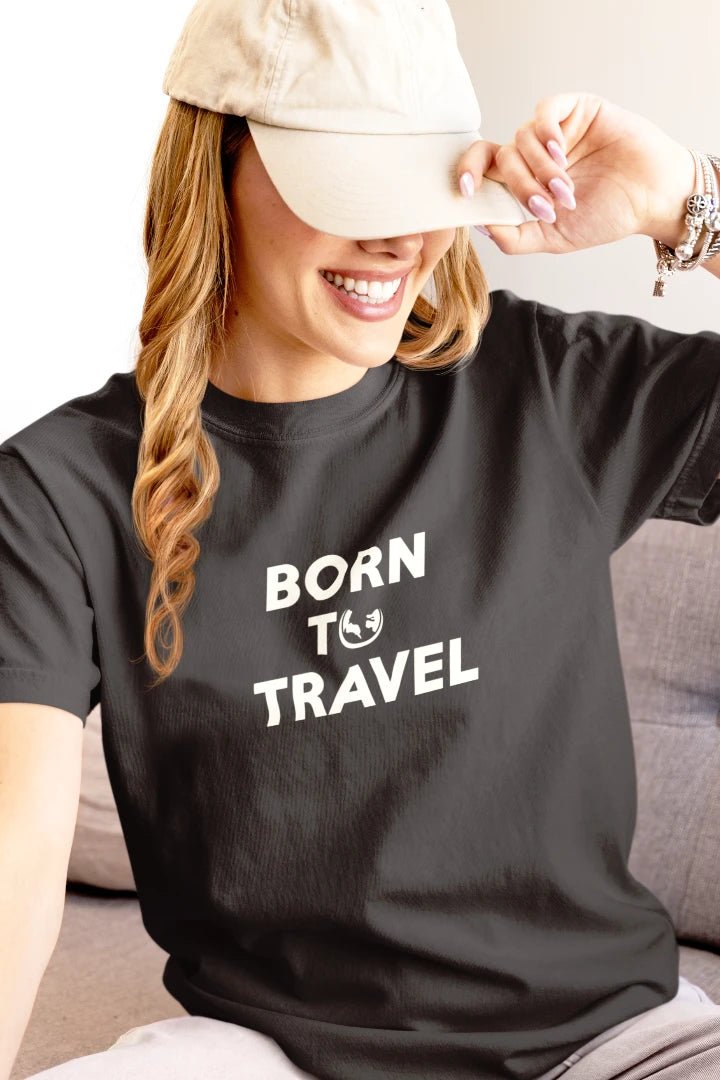 A woman wearing a Graphite crewneck t-shirt with the white text 'BORN TO TRAVEL' and a beige baseball cap, sitting on a gray couch.