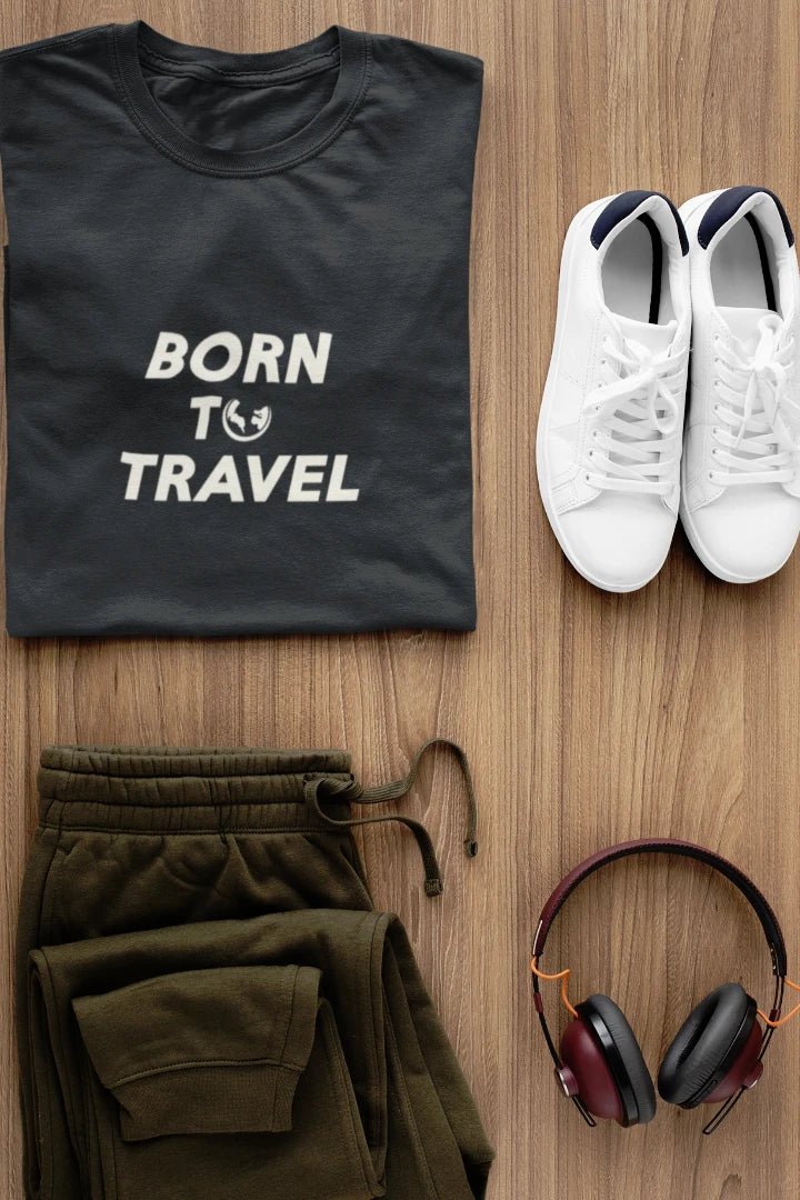 A styled flat-lay featuring a Graphite t-shirt with the white text 'BORN TO TRAVEL' and a small globe graphic, paired with olive green sweatpants, white sneakers, and over-ear headphones on a light brown wood background.