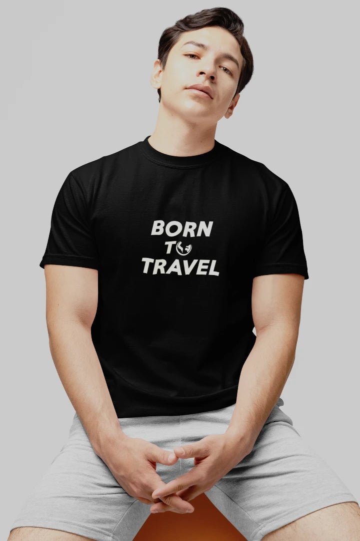 A man wearing a Jet black crewneck t-shirt with the white printed text 'BORN TO TRAVEL' and light gray shorts, seated against a solid gray background.
