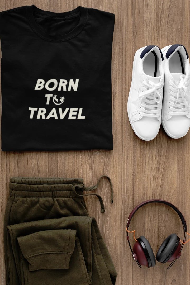 A styled flat-lay of a Jet black t-shirt with the white text 'BORN TO TRAVEL' and a small globe graphic, paired with olive green sweatpants, white sneakers, and over-ear headphones on a light brown wood background.