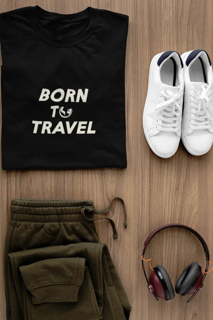 A styled flat-lay of a Jet black t-shirt with the white text 'BORN TO TRAVEL' and a small globe graphic, paired with olive green sweatpants, white sneakers, and over-ear headphones on a light brown wood background.
