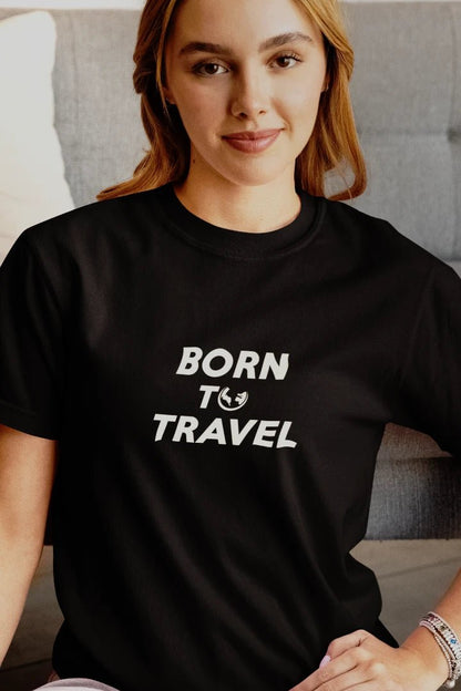 A woman wearing a Jet black short-sleeve t-shirt with the text 'BORN TO TRAVEL' printed in white on the front, posed in front of a gray couch.