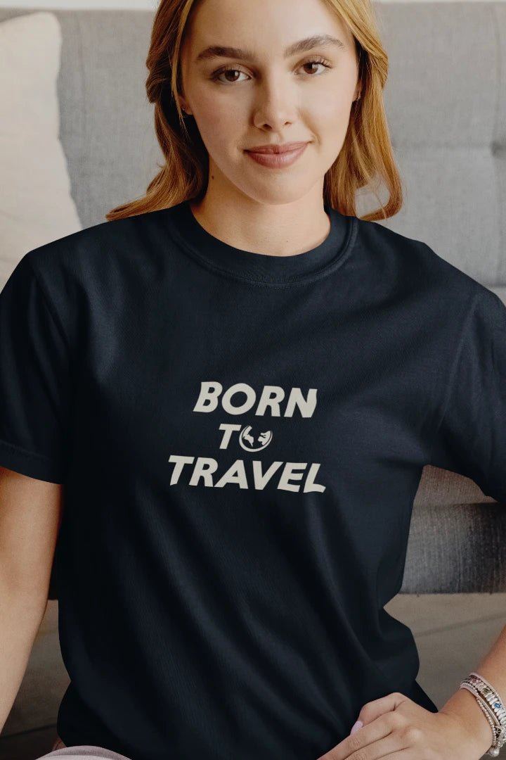 A woman wearing a Midnight Navy crewneck t-shirt with the text 'BORN TO TRAVEL' printed in white across the chest, sitting in a cozy indoor setting.