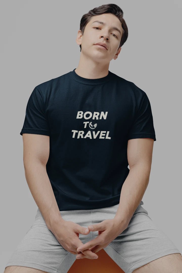 A man wearing a dark Midnight Navy t-shirt with the text 'BORN TO TRAVEL' printed in white across the chest, paired with light gray shorts, seated against a solid gray background.