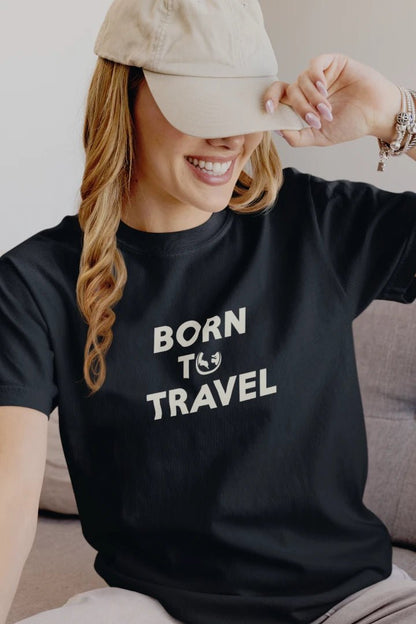 A woman wearing a Midnight Navy crewneck t-shirt with the text 'BORN TO TRAVEL' in white, paired with a beige baseball cap, sitting on a couch.