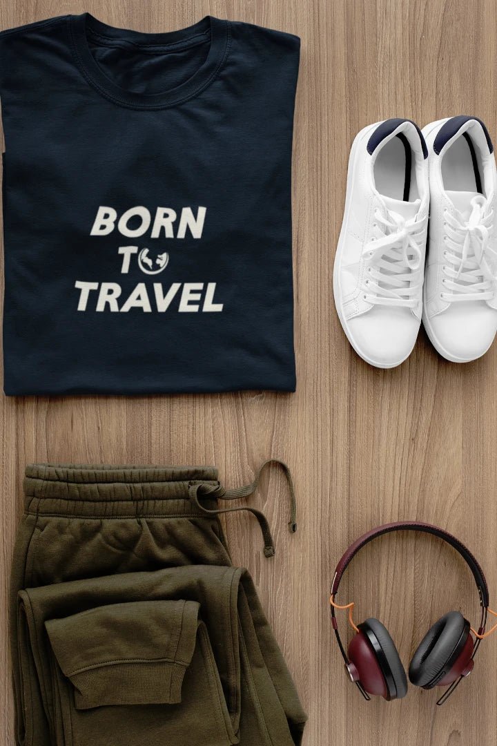 A styled flat-lay outfit on a light wood background featuring a Midnight Navy t-shirt with 'BORN TO TRAVEL' printed with sweatpants, white sneakers, and burgundy over-ear headphones.