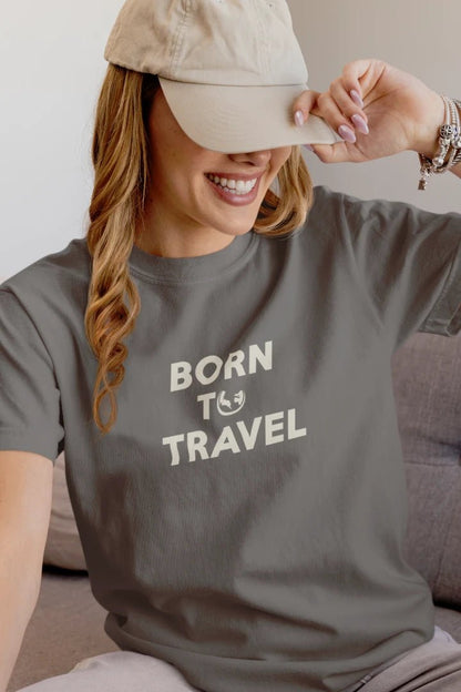 A woman wearing a dark Heather Charcoal t-shirt with the text 'BORN TO TRAVEL' in white, paired with a beige baseball cap, sitting on a couch.
