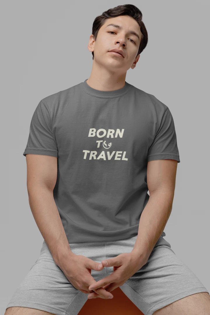 A man wearing a dark Heather Charcoal crewneck t-shirt with the text 'BORN TO TRAVEL' printed in cream on the chest, paired with light gray shorts, set against a solid gray background.