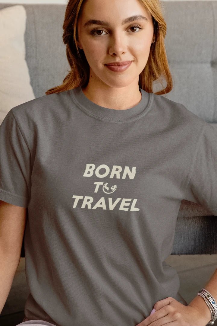A woman wearing a dark Heather Charcoal crewneck t-shirt with the text 'BORN TO TRAVEL' printed in cream on the chest, sitting in front of a gray tufted sofa.