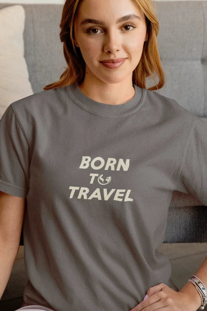 A woman wearing a dark Heather Charcoal crewneck t-shirt with the text 'BORN TO TRAVEL' printed in cream on the chest, sitting in front of a gray tufted sofa.