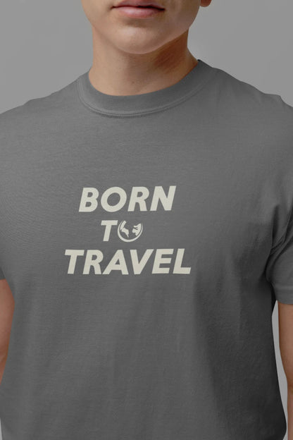 A close-up of a person wearing a dark Heather Charcoal crewneck t-shirt with the text 'BORN TO TRAVEL' and a small globe graphic printed in off-white on the chest, set against a solid gray background.