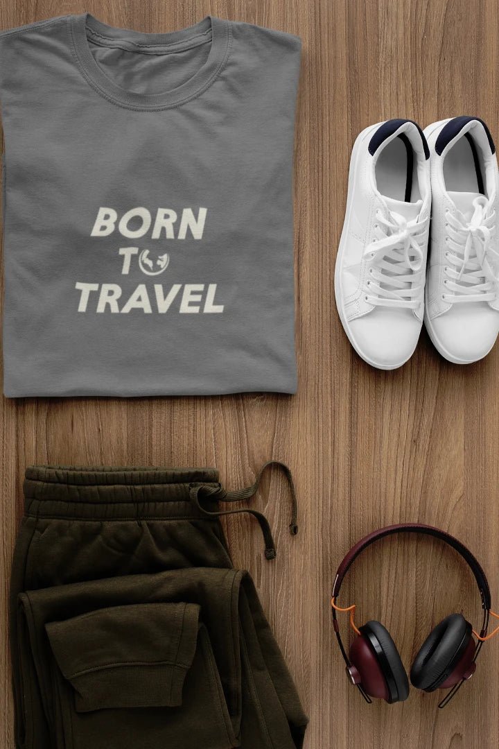 A styled flat-lay featuring a dark Heather Charcoal t-shirt with 'BORN TO TRAVEL' printed in white, olive green sweatpants, white sneakers, and  headphones on a light brown wood background.