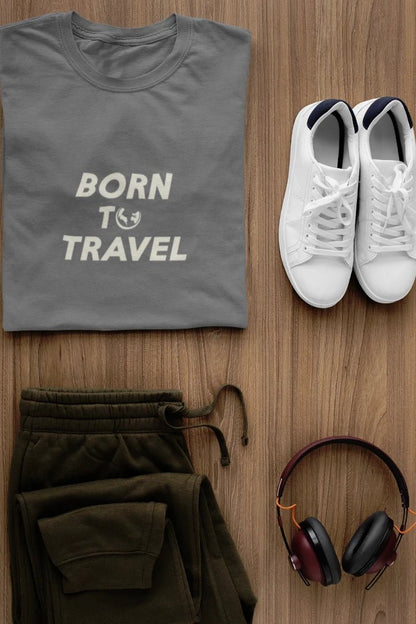 A styled flat-lay featuring a dark Heather Charcoal t-shirt with 'BORN TO TRAVEL' printed in white, olive green sweatpants, white sneakers, and  headphones on a light brown wood background.