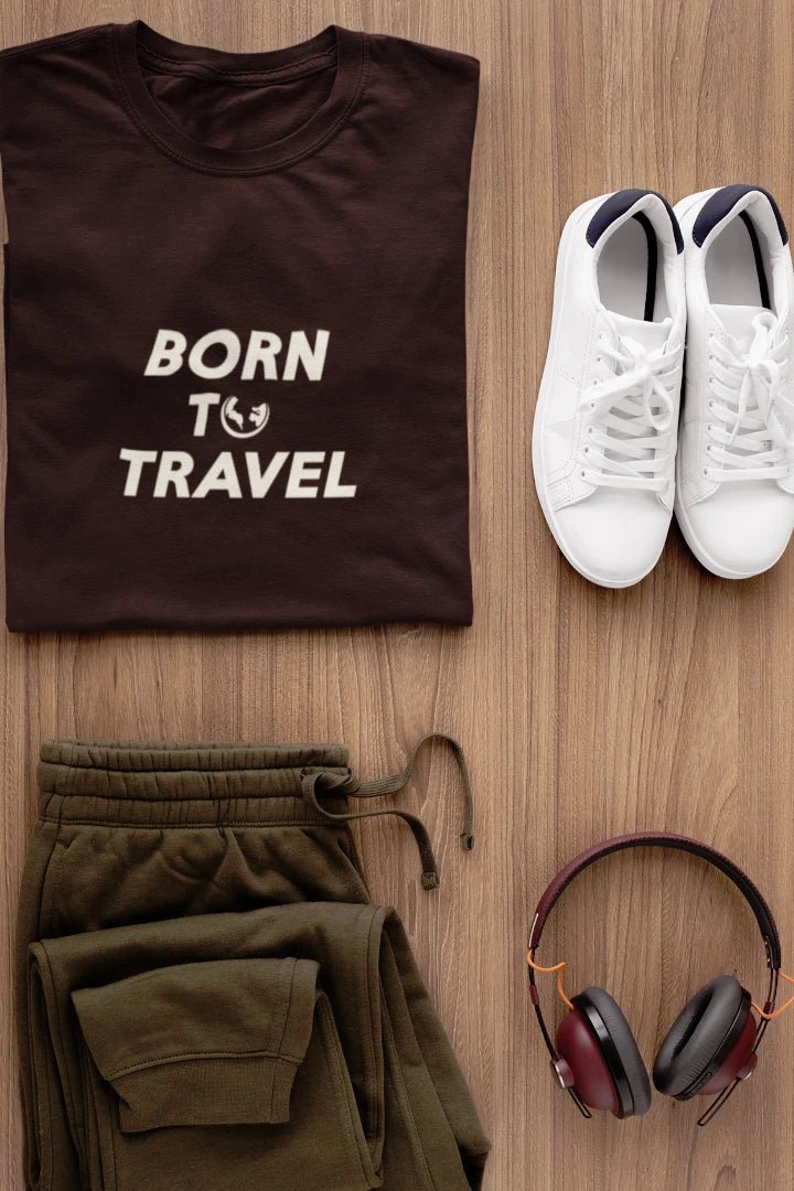 A styled flat-lay of a Dark Mocha t-shirt with the text 'BORN TO TRAVEL' in white, paired with olive green sweatpants, white sneakers, and over-ear headphones on a light wood background.