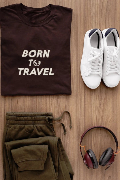 A styled flat-lay of a Dark Mocha t-shirt with the text 'BORN TO TRAVEL' in white, paired with olive green sweatpants, white sneakers, and over-ear headphones on a light wood background.