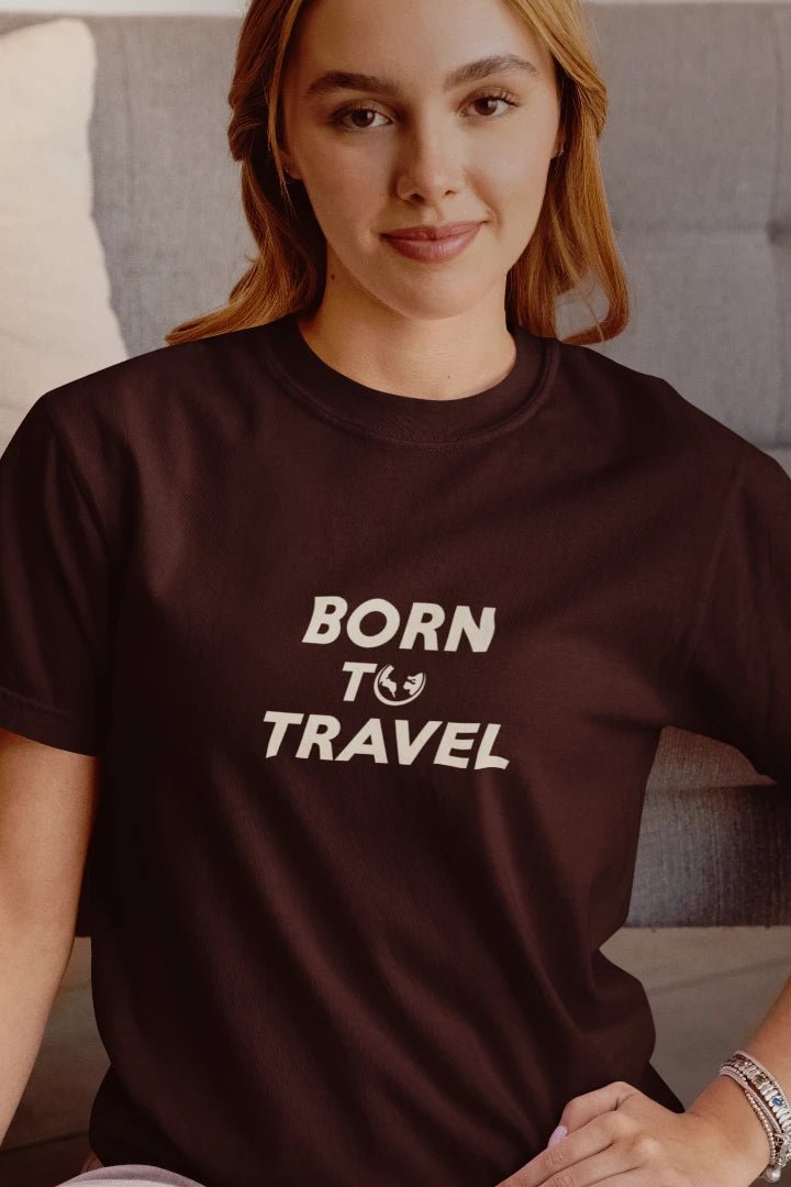 A woman wearing a Dark Mocha crewneck t-shirt with the text 'BORN TO TRAVEL' printed in white on the chest, sitting in front of a gray couch.