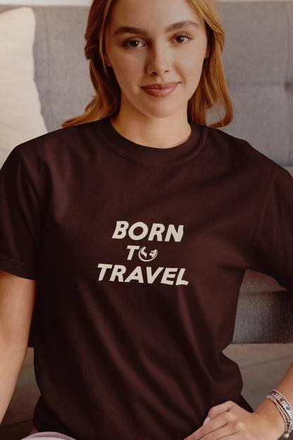 A woman wearing a Dark Mocha crewneck t-shirt with the text 'BORN TO TRAVEL' printed in white on the chest, sitting in front of a gray couch.
