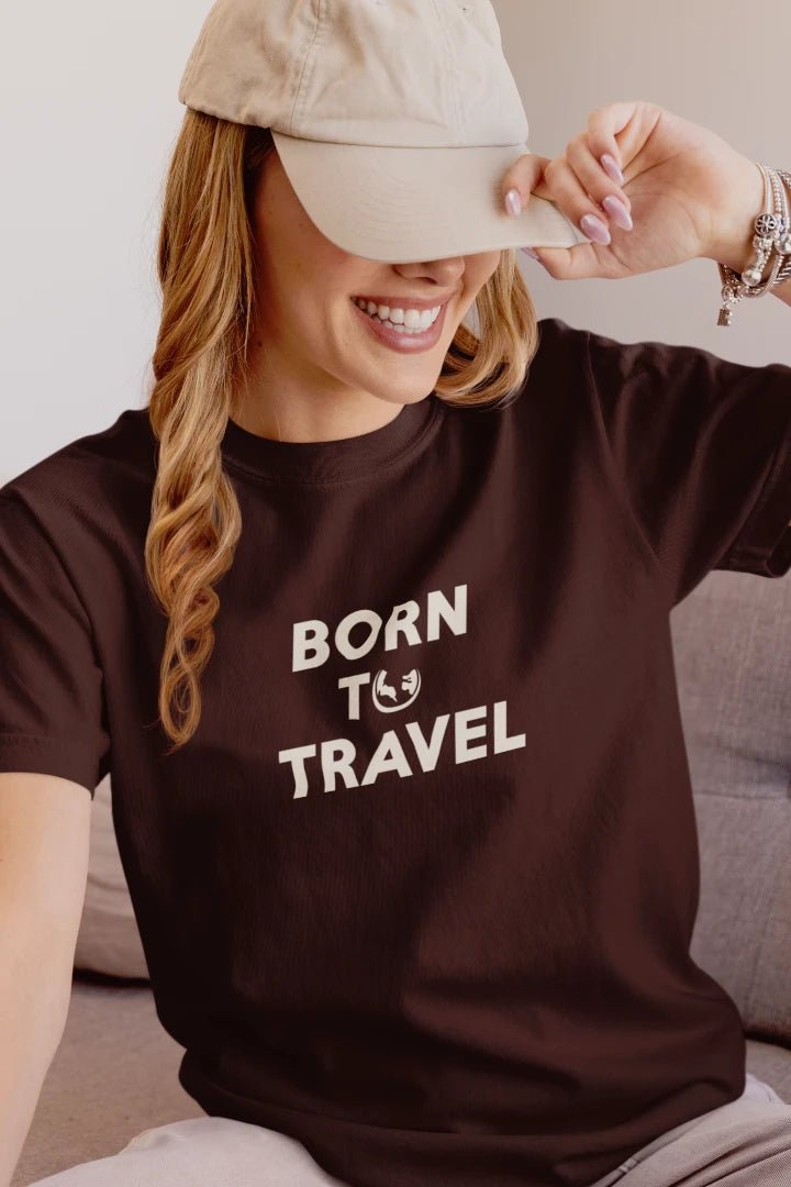 A woman wearing a Dark Mocha t-shirt with the text 'BORN TO TRAVEL' in white, sitting on a couch and holding a beige baseball cap.