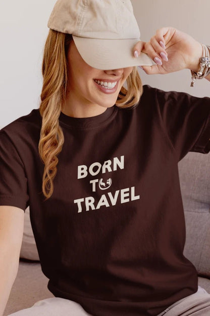 A woman wearing a Dark Mocha t-shirt with the text 'BORN TO TRAVEL' in white, sitting on a couch and holding a beige baseball cap.
