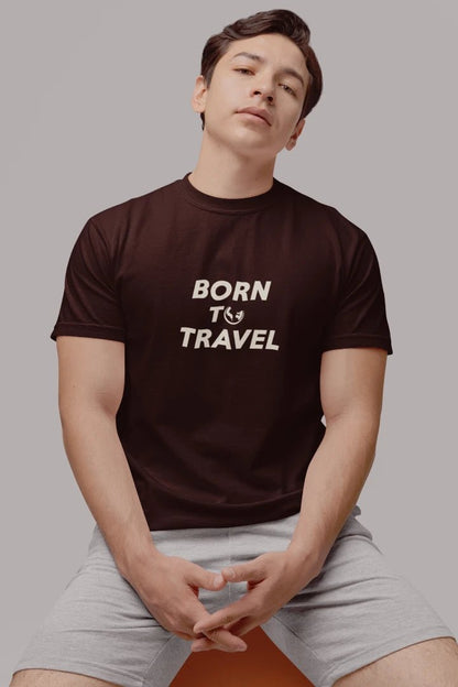 A man wearing a Dark Mocha crewneck t-shirt with the text 'BORN TO TRAVEL' printed in white on the chest, paired with light gray shorts, seated against a solid gray background.