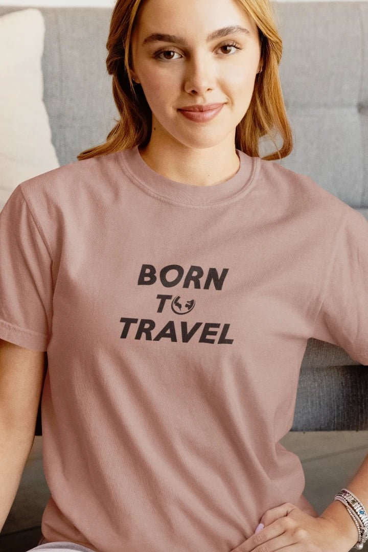 A woman wearing a dusty Taupe t-shirt with the text 'BORN TO TRAVEL' printed in black on the chest, sitting in front of a gray couch.
