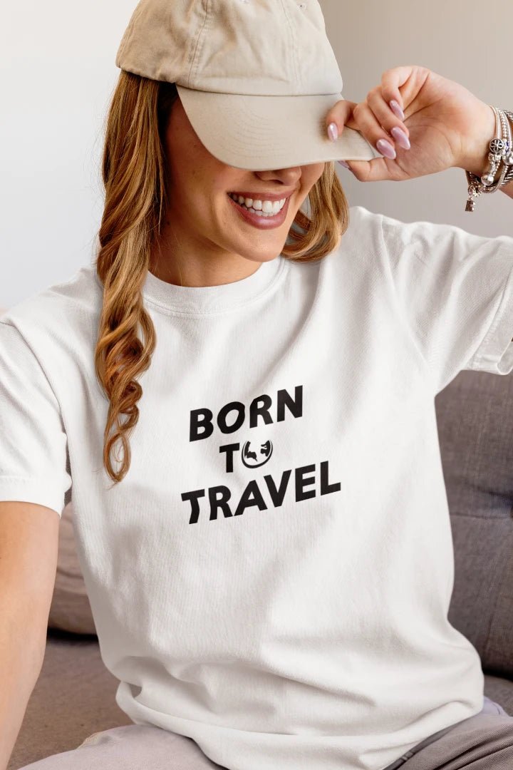 A woman wearing a Pure White crewneck t-shirt with the black printed text 'BORN TO TRAVEL' and a small smiley face graphic, paired with a beige baseball cap.